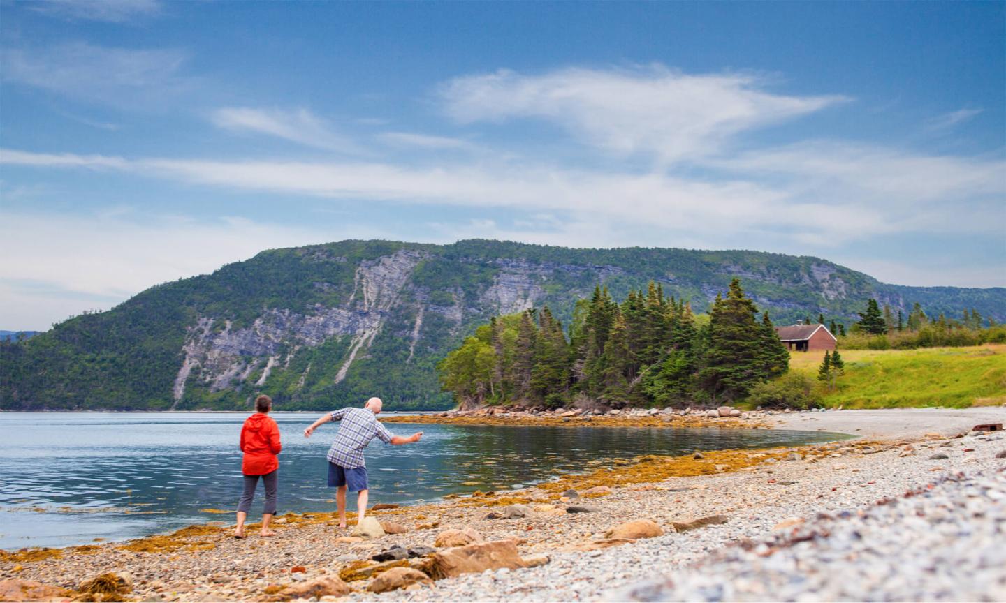 Two people skipping stones on a rocky beach with a forested mountain in the background.