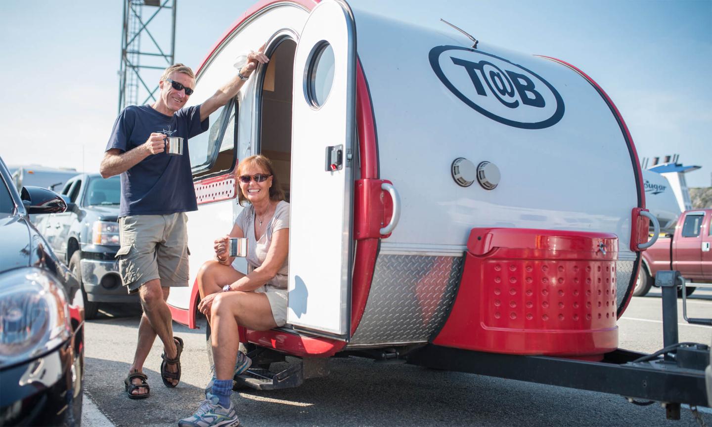 A smiling couple enjoying coffee outside their red and white camper trailer.
